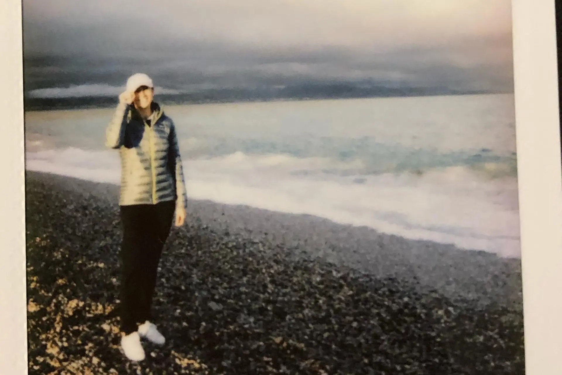 Bord de mer avec des galets et un humain debout, casquette sur la tête, sourire et visière de la casquette dans la main